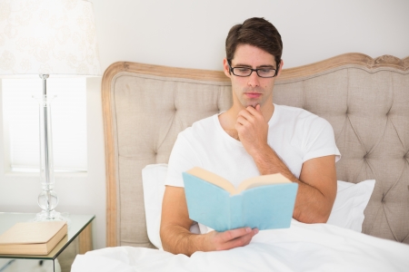 Relaxed young man reading book in bed at houseの写真素材