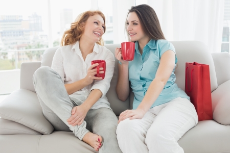 Two happy young female friends with coffee cups conversing in the living room at homeの写真素材