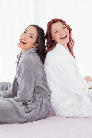 Side view portrait of two cheerful young female friends in bathrobes sitting back to back on bed at homeの写真素材