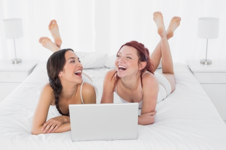 Relaxed cheerful female friends with laptop lying in bed at homeの写真素材