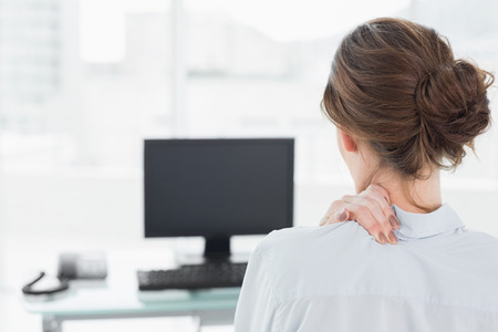 Rear view of a businesswoman with neck pain in front of computer in a bright officeの写真素材