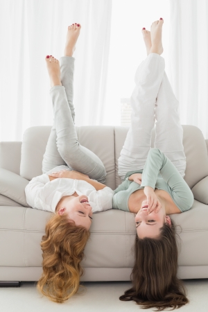 Two smiling young female friends lying on sofa with legs in the air in living room at homeの写真素材