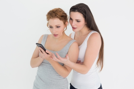 Two casual young female friends looking at mobile phone against white backgroundの写真素材