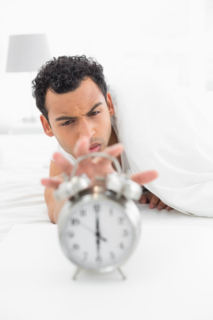 Close-up of a sleepy young man in bed extending hand to alarm clock at homeの写真素材