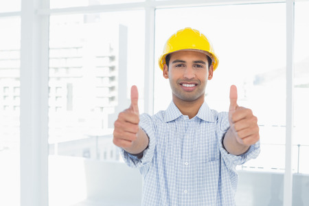 Portrait of a smiling handyman in yellow hard hat gesturing thumbs up in a bright officeの写真素材