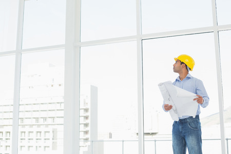 Young architect in yellow hard hat with plans looking up at window in a bright officeの写真素材