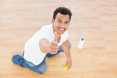Portrait of a smiling young man cleaning the floor while gesturing thumbs up at houseの写真素材