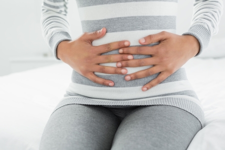 Close-up mid section of a casual young woman with stomach pain sitting in bed at homeの写真素材