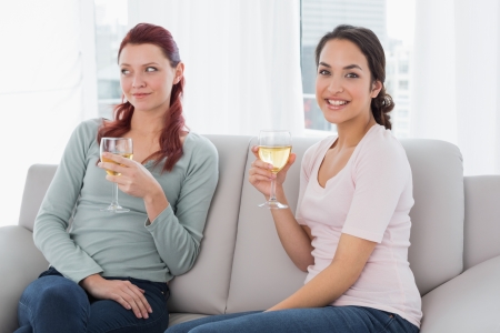 Two young female friends with wine glasses sitting on sofa at homeの写真素材