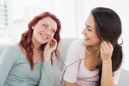 Two cheerful young female friends listening music through earphones together in the living room at homeの写真素材