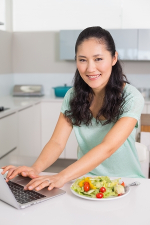 Portrait of a smiling young woman using laptop while having salad in the kitchen at homeの写真素材