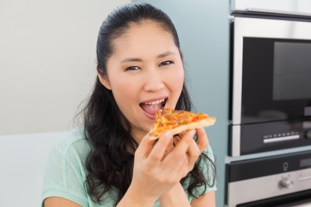 Close-up portrait of a smiling young woman eating a slice of pizza in the kitchen at homeの写真素材