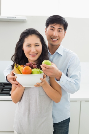 Portrait of a young man and woman holding bowl full of fruit in the kitchen at homeの写真素材