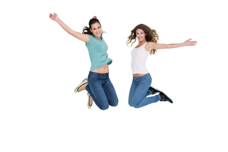 Full length side view of two cheerful young female friends jumping over white background