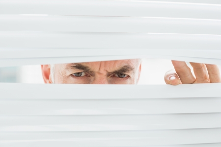Close-up portrait of a serious mature businessman peeking through blinds in the officeの写真素材