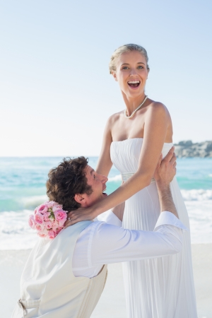 Laughing groom picking up his pretty blonde wife smiling at camera at the beachの写真素材
