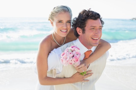 Handsome groom giving his pretty wife a piggy back at the beachの写真素材
