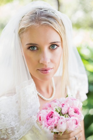Happy bride wearing veil holding bouquet looking at camera in the countrysideの写真素材