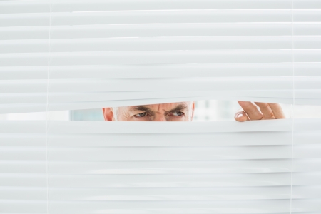 Close-up of a serious mature businessman peeking through blinds in the officeの写真素材