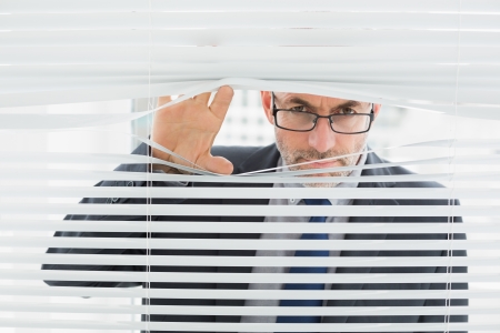 Close-up portrait of a serious mature businessman peeking through blinds in the officeの写真素材