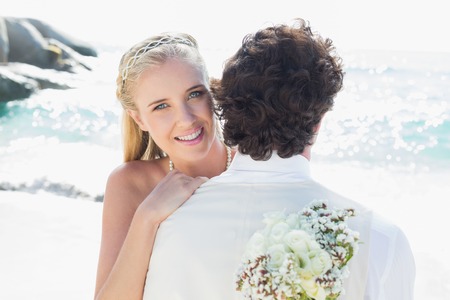 Pretty blonde bride smiling at camera while hugging husband at the beachの写真素材