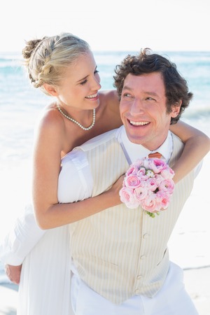 Happy groom giving his blonde wife a piggy back at the beachの写真素材