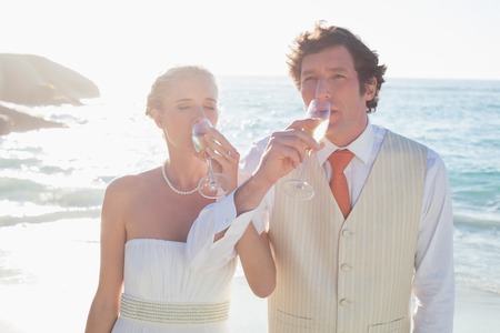 Young newlyweds drinking champagne linking arms at the beachの写真素材