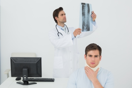 Patient in surgical collar with male doctor examining spine x-ray in background at medical officeの写真素材