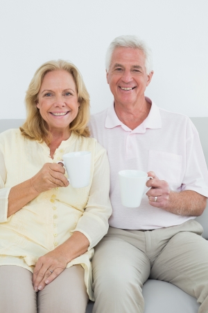 Relaxed senior couple with coffee cups sitting on sofa in a houseの写真素材