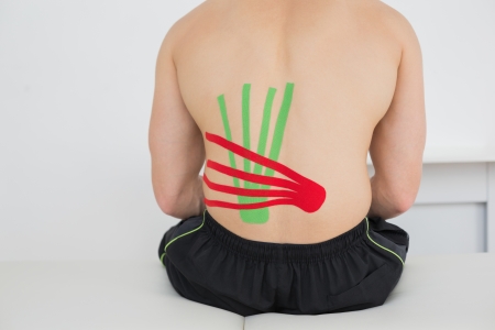 Rear view of a shirtless young man with red and green kinesio tapes on back in the medical office