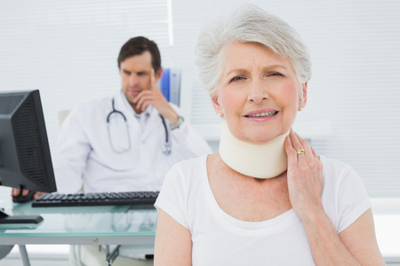 Senior patient in surgical collar with male doctor sitting at desk in background at medical officeの写真素材