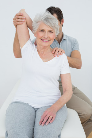 Male physiotherapist stretching a smiling senior woman's arm in the medical officeの写真素材