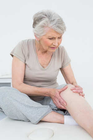 Senior woman with her hands on a painful knee while sitting on examination tableの写真素材