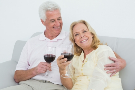 Relaxed senior couple with wine glasses sitting on sofa at homeの写真素材