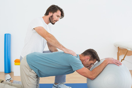 Physical therapist assisting young man with yoga ball in the gym at hospitalの写真素材