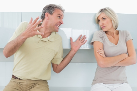 Tired woman sitting with arms crossed as man argue in the kitchen at homeの写真素材