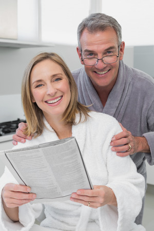Happy couple in bathrobes reading newspaper in the kitchen at homeの写真素材