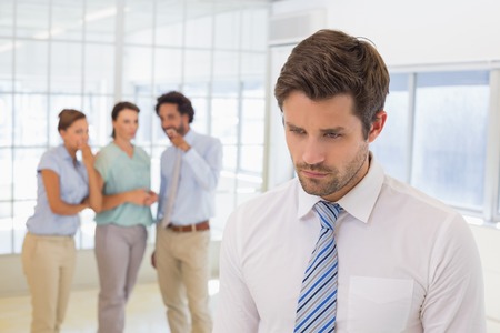 Colleagues gossiping with sad young businessman in foreground at a bright officeの写真素材