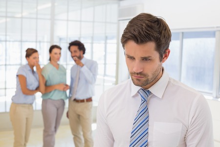 Colleagues gossiping with sad young businessman in foreground at a bright office