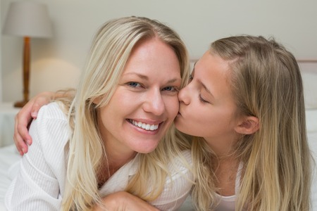 Cute girl kissing mother on cheek in bedの写真素材