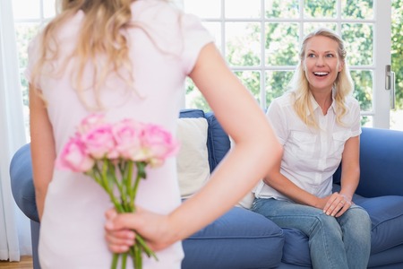 Happy mother looking at girl hiding flower bouquet behind back at homeの写真素材
