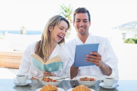 Cheerful young couple with book and digital tablet on breakfast table at homeの写真素材