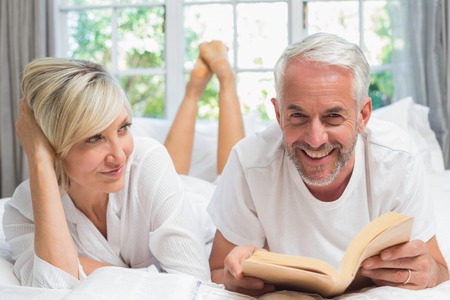 Happy mature couple lying and reading book in bed at homeの写真素材