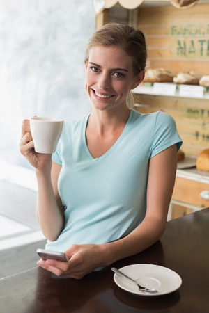 Smiling young woman drinking coffee while using mobile phone at counter in the coffee shopの写真素材