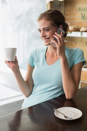 Smiling young woman drinking coffee while using mobile phone at counter in the coffee shopの写真素材