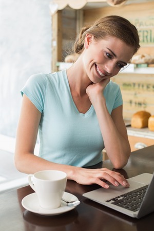 Smiling young woman with coffee cup using laptop at counter in the coffee shopの写真素材