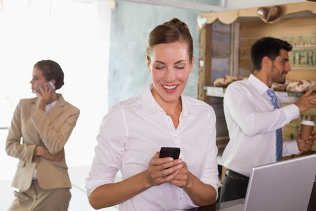 Businesswoman using mobile phone and laptop with colleagues behind in office cafeteriaの写真素材