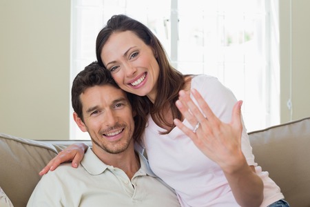Portrait of a happy young woman showing engagement ring besides man at homeの写真素材