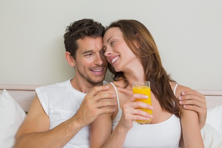 Happy relaxed young couple with orange juice in bed at homeの写真素材