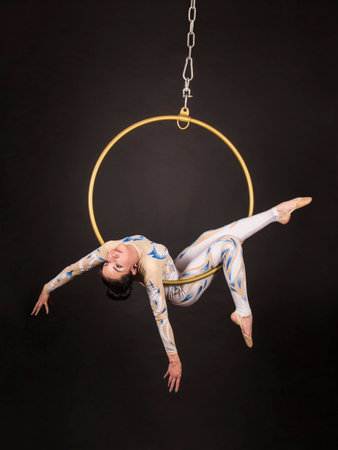 A slender,dark-haired girl - an air acrobat in a white and blue suit, performs exercises in an air ring. Studio shooting on a dark background.の写真素材